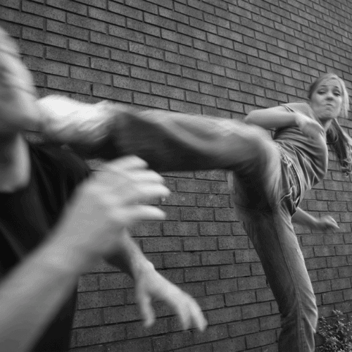 A person performs a high kick toward another person's head against a brick wall background.